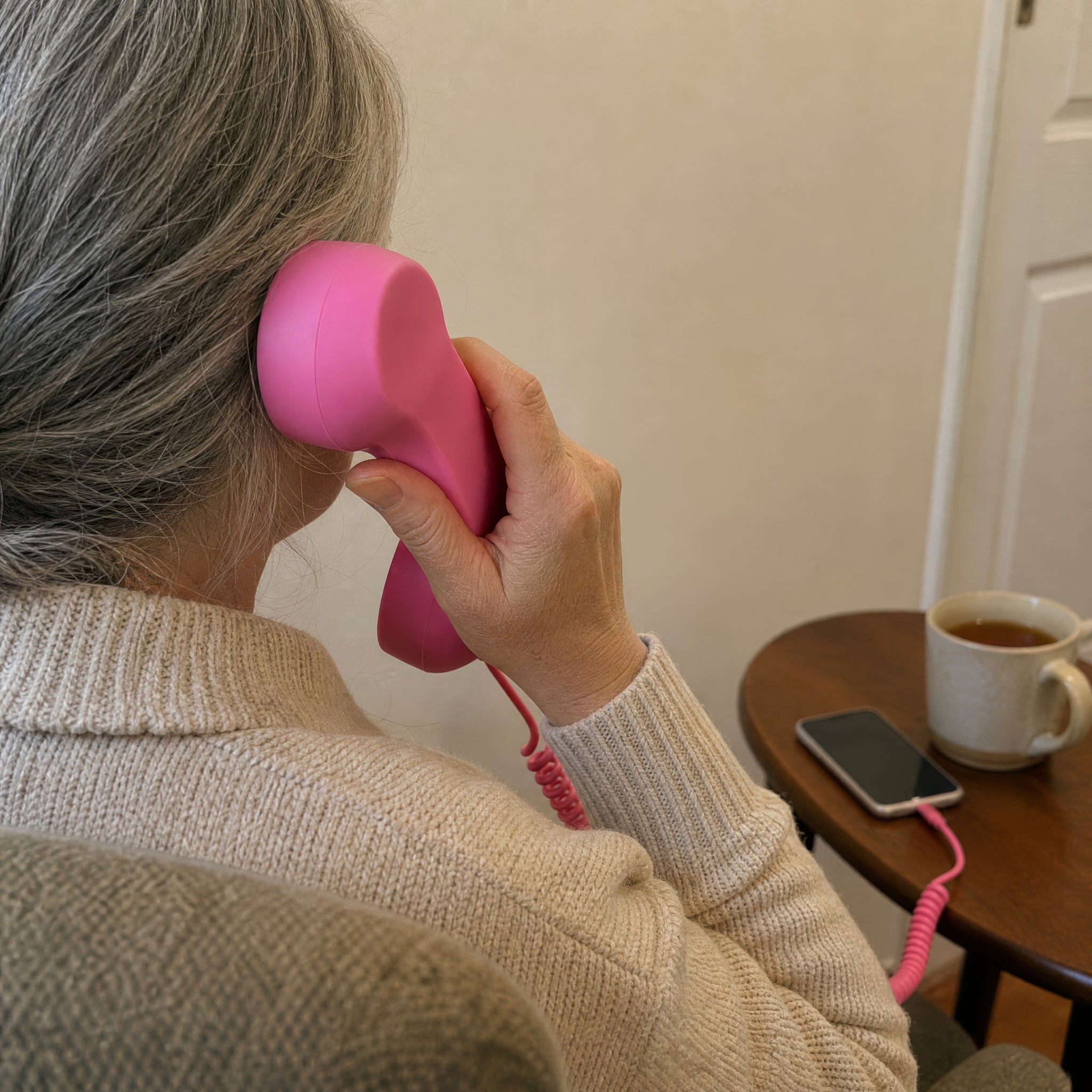A woman in an armchair holds the pink DialBoop handset to her ear, cord running to her phone on the side table